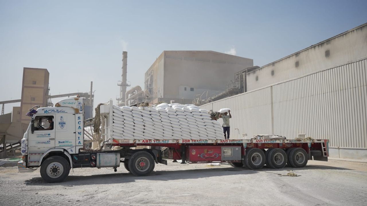 Truck loaded with white sacks parked near a large industrial building under a clear sky.